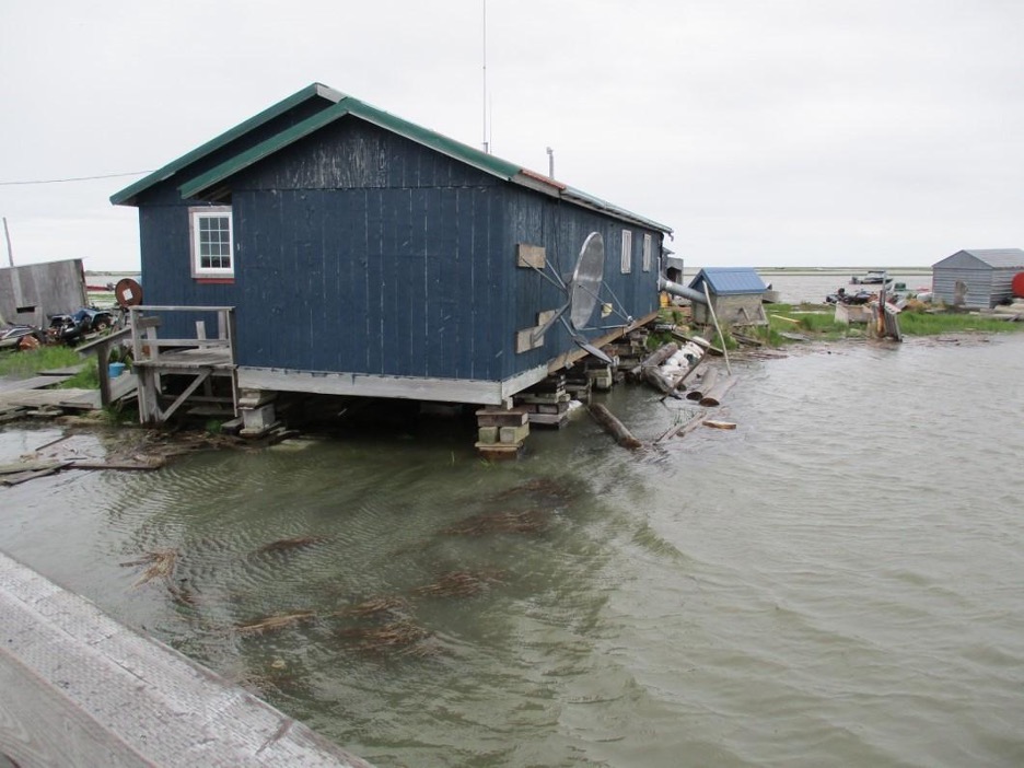 Image of blue house elevated on blocks and surrounded by floodwaters