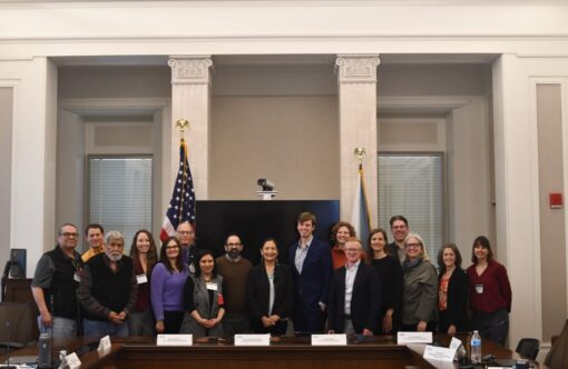 16 members of the Advisory Council for Climate Adaptation Science stand for photo with Sec. Deb Haaland (center) behind meeting table and before two flags, the US and Dept. of Interior.