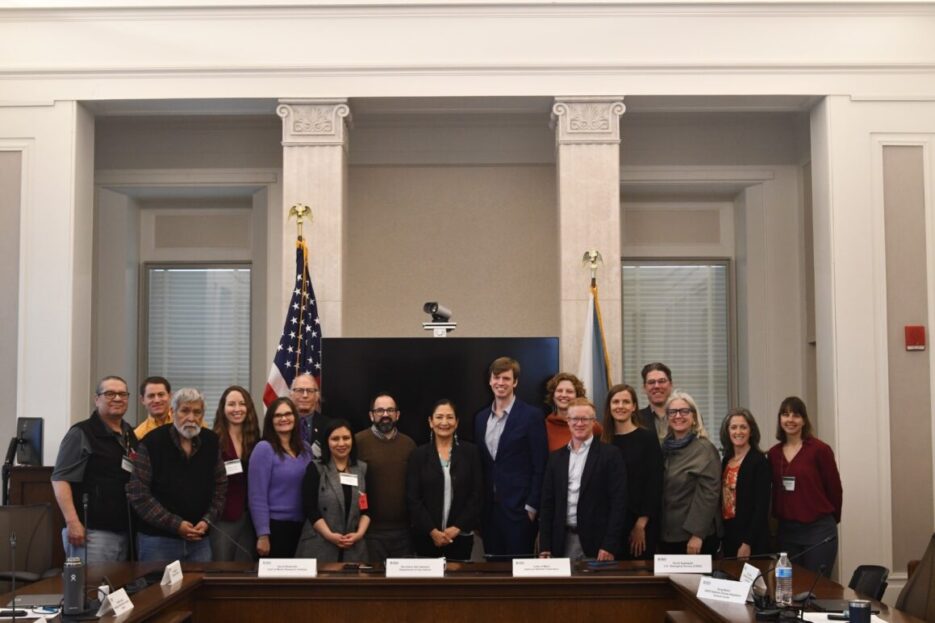 16 members of the Advisory Council for Climate Adaptation Science stand for photo with Sec. Deb Haaland (center) behind meeting table and before two flags, the US and Dept. of Interior.